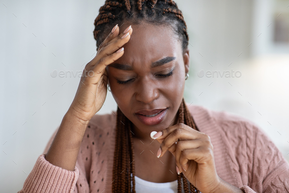 Upset black lady suffering from headache or migraine at home, taking pill