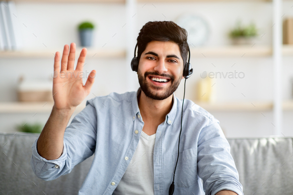 Greeting concept. Arab man wearing headset and waving hand to camera, gesturing hello and smiling