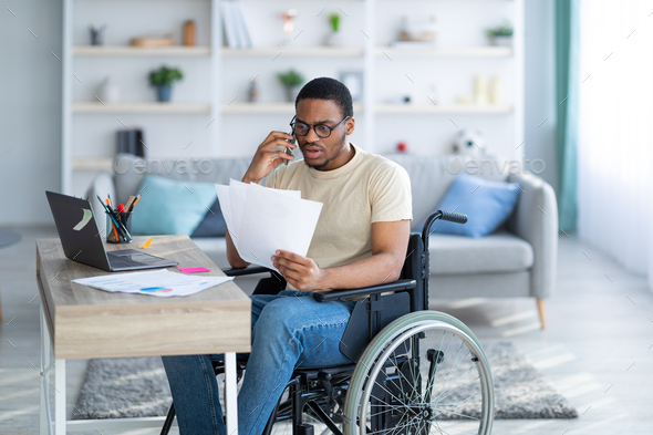 Online work for disabled people. Young black guy with documents speaking on phone in front of laptop