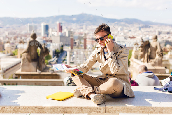Enjoyed guy in sunglasses is sitting on sunny city background. He wears beige coat, speaking on