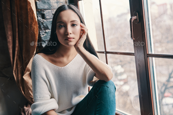 No worries. Attractive young woman looking at camera while sitting on the window sill at home