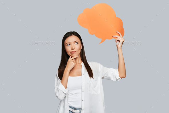 What if? Beautiful young Asian woman keeping hand on chin while standing against grey background