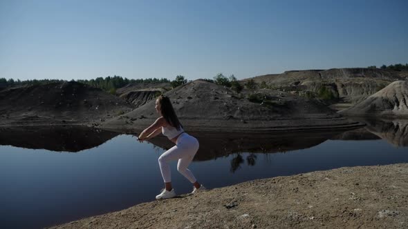Woman in a White Tracksuit Performs Squats with Lunges on the Shore of the Lake