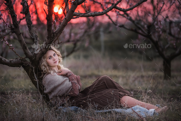 Young beautiful curly blonde woman sits in blooming peach sunset gardens