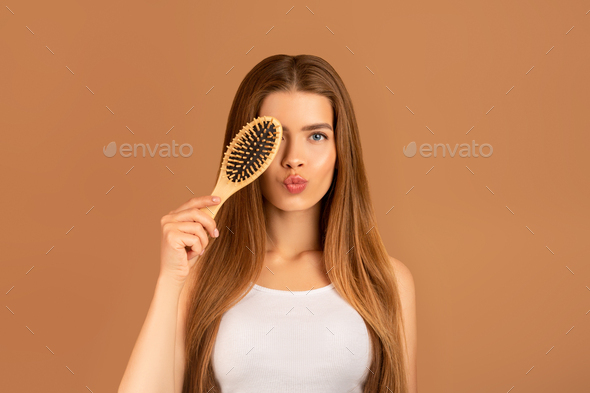 Natural cosmetics for silky hair. Portrait of lovely young lady holding hairbrush near her eye on