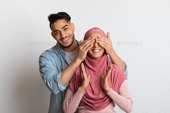 Loving arab man surprising his excited wife in hijab, covering her eyes