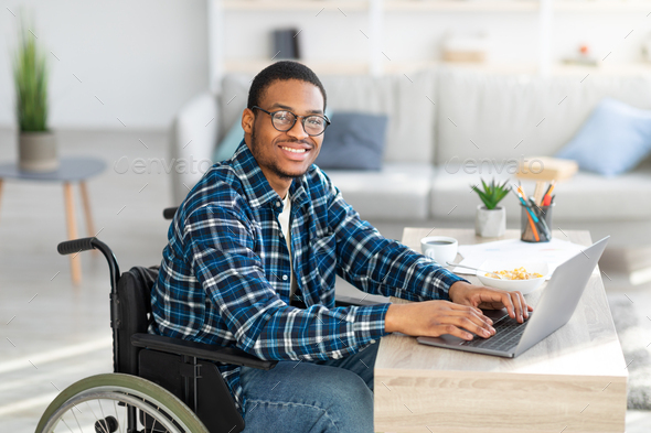 Cheerful handicapped black man in wheelchair using laptop for online job from home, having business