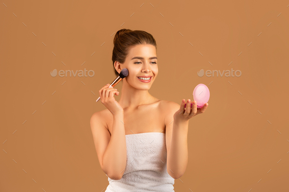 Charming young lady using cosmetic brush to apply decorative makeup, looking in mirror on brown