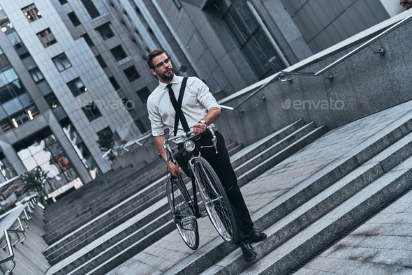 Just in time. Good looking young man in full suit carrying his bicycle while walking outdoors