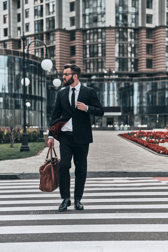 Busy outdoors. Full length of young man in full suit crossing the street  while walking outdoors Stock Photo by gstockstudio