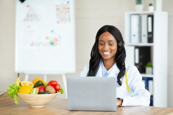 Cheerful black female dietitian using laptop pc, creating online weight loss program at modern