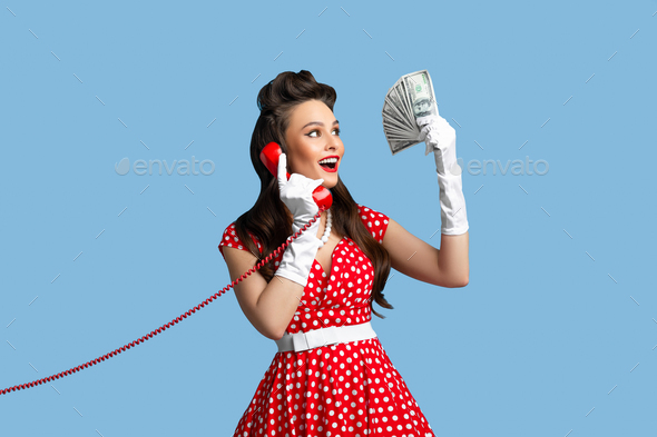 Happy pin up woman in cool polka dot red dress holding fan of money and speaking on landline phone