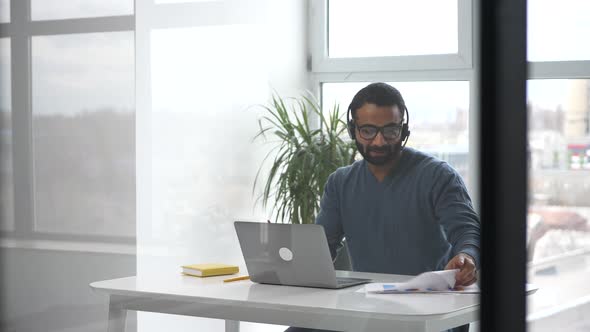 Handsome Young Indian Man Wearing Headset and Stylish Glasses Using Laptop in the Office alt