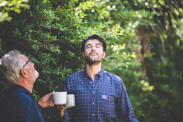 An adult hipster son and senior father drinking coffee in the garden at home