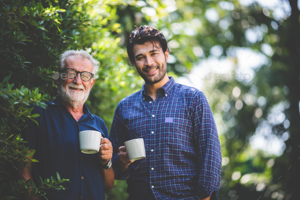 An adult hipster son and senior father drinking coffee in the garden at home