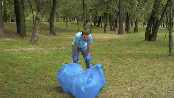 Wide Shot Portrait of Exhausted Caucasian Man Standing in Park with Garbage Bags and Wiping Forehead alt