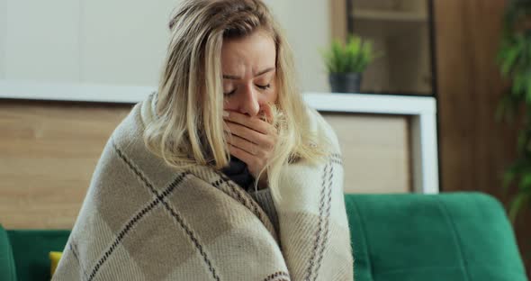 Sick Young Woman at Home on the Sofa with a Cold She is Covering with a Blanket and Blowing Her Nose alt
