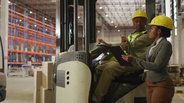 African american male and female workers wearing safety suits and talking in warehouse alt