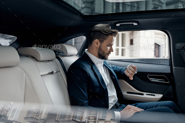 Just in time. Handsome young man in full suit looking at his watch while sitting in the car