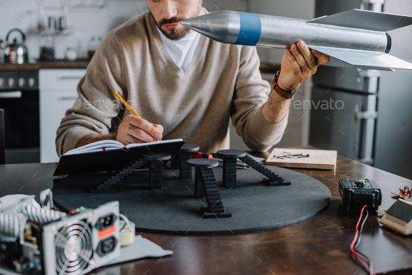 cropped image of engineer holding rocket model and making notes at home