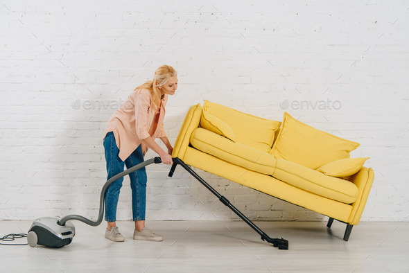 Full length view of senior woman with vacuum cleaner cleaning floor under yellow sofa