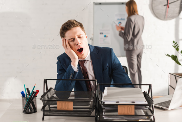 selective focus of tired man yawning near document trays with lettering with female coworker on