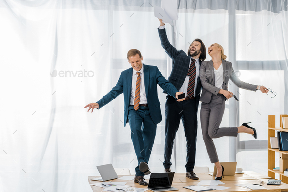 joyful insurance workers dancing on table and throwing papers at meeting in office