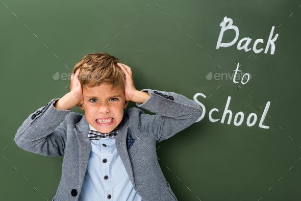 angry schoolboy holding his head next to chalkboard with back to school sign