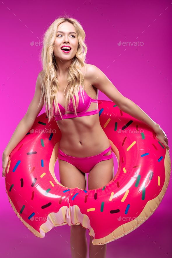 Cheerful blonde woman in bikini wearing swimming tube in shape of doughnut and looking away