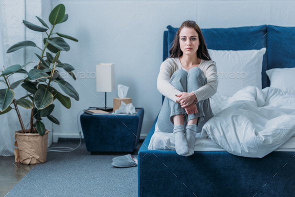 upset woman hugging knees, looking at camera and sitting on bed at home
