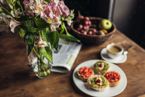 Still life of newspaper, flowers and breakfast with cakes and hot coffee on kitchen table in front