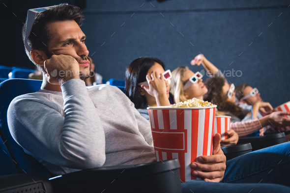bored multiethnic friends with popcorn watching film together in movie theater