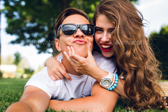 Selfie-portrait of funny couple lying on grass in summer park. Girl with long curly hair, red lips a - Stock Photo - Images