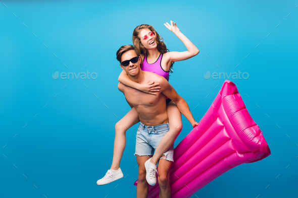 Cute girl with long curly hair is riding on back of hot guy with naked torso on blue background in s