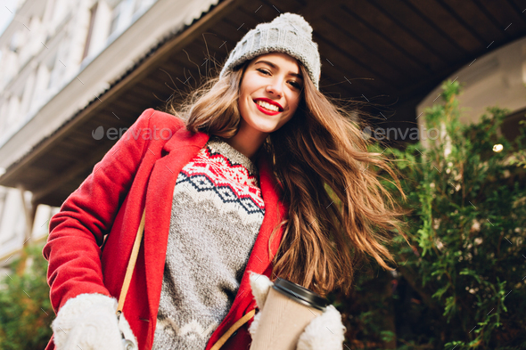 Pretty girl with long hair in knitted hat, red coat walking on street with coffee to go. She wears w