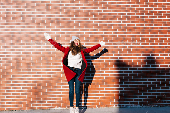 Pretty young girl with long hair in red coat and white gloves on wall background outside. She is jum
