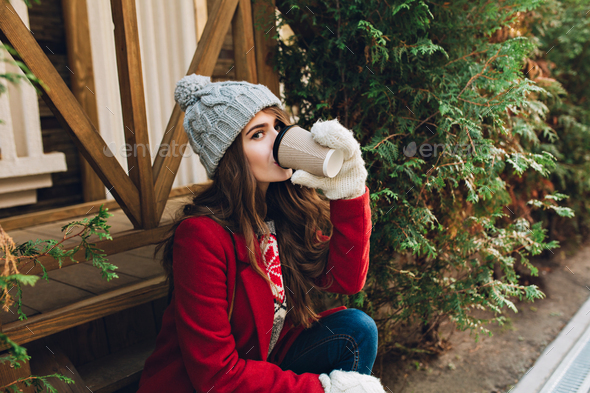 Portrait pretty girl with long hair in red coat, knitted hat and white gloves sitting on wooden stai