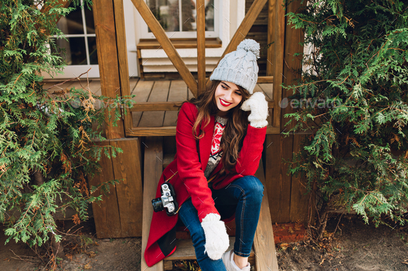 Pretty brunette girl in red coat, knitted hat and white gloves sitting on wooden stairs outdoor. She