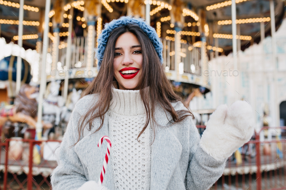 Portrait of blissful long-haired female model in white gloves holding candy cane in amusement park.