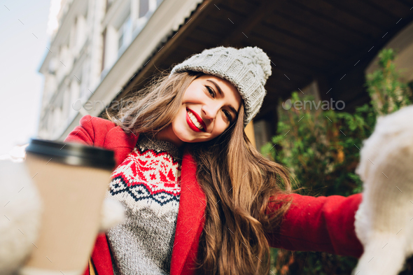 Selfie portrait pretty girl with long hair in red coat on street. She wears knitted hat, white glove