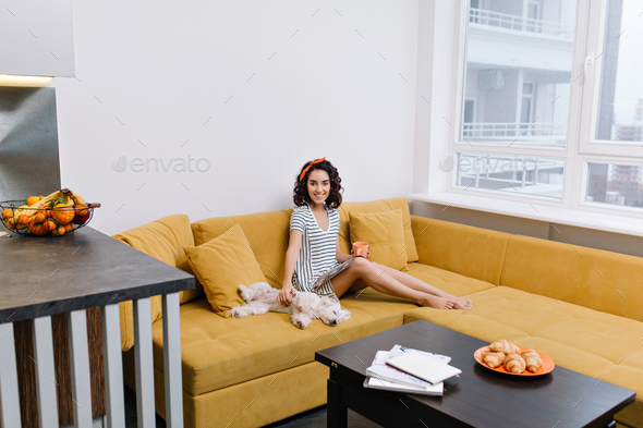 Relax time in modern apartment of enjoyed, happy young woman chilling on orange couch. Magazine, cup