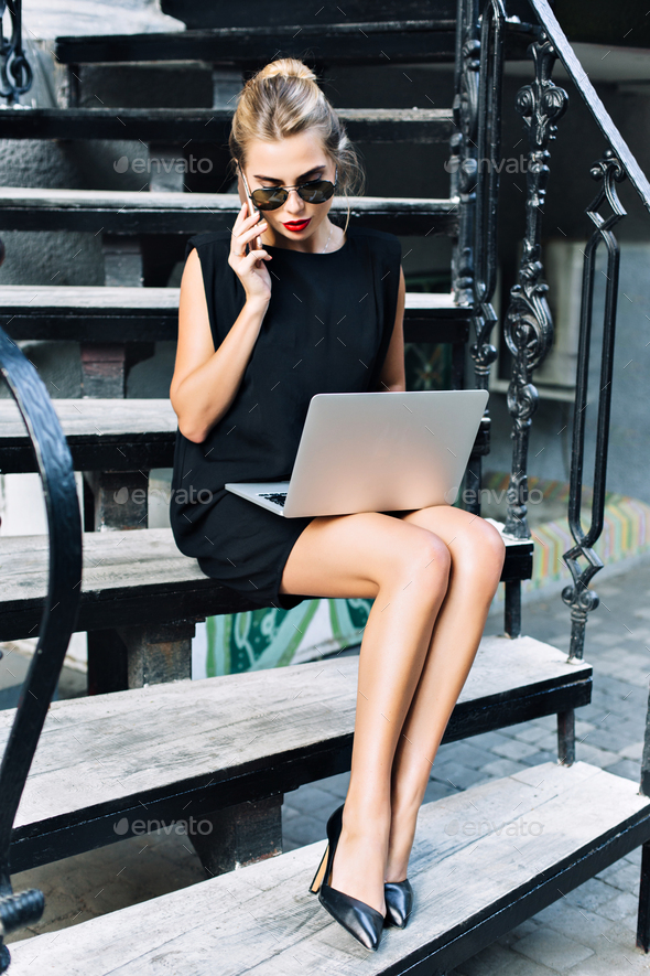 Pretty businesswoman in black short dress sitting on stairs outdoor. She wears high heels, working w