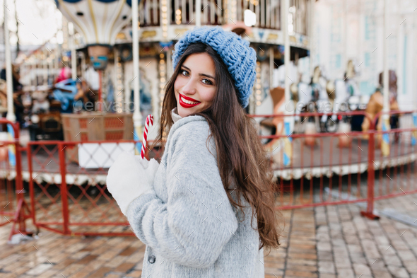 Portrait of blissful long-haired female model in white gloves holding candy cane in amusement park.