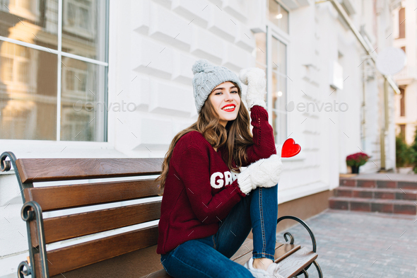 Full-length young girl with long hair in knitted hat, jeans and white gloves sitting on bench on str