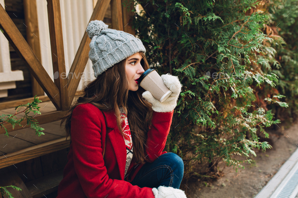 Portrait beautiful girl with long hair in red coat, knitted hat and white gloves sitting on wooden s