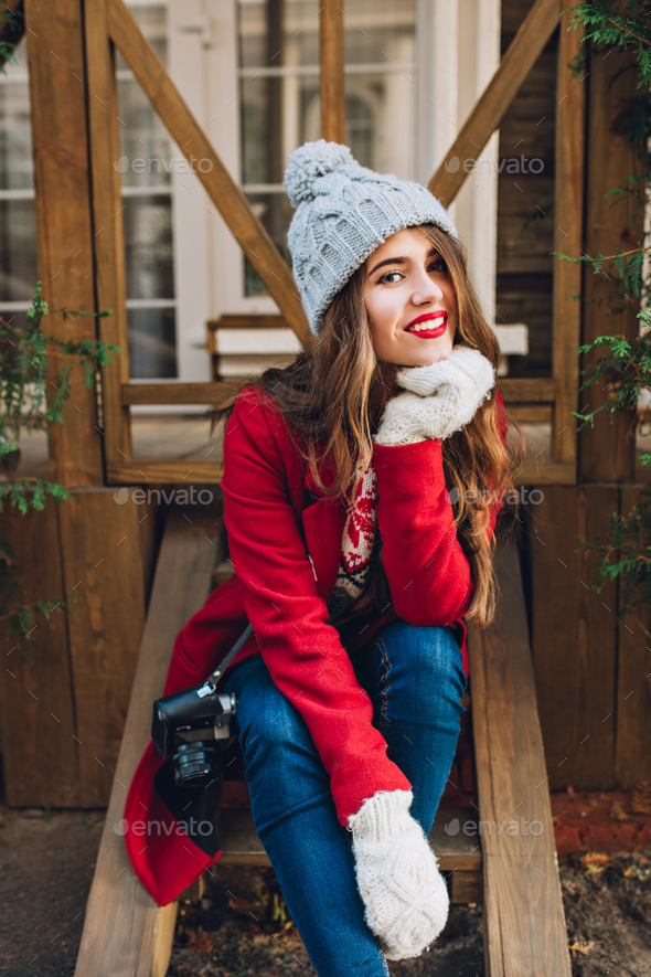 Pretty brunette girl in red coat, knitted hat and white gloves sitting on wooden stairs outdoor. She