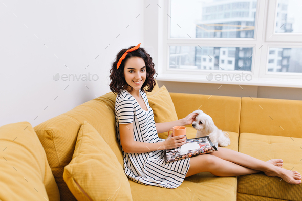 Joyful young woman with cut brunette hair in dress chilling with dog on couch in modern apartment. R