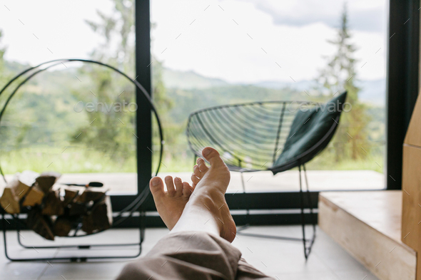 Beautiful female feet on background of firewood and big window in modern chalet, woman relaxing