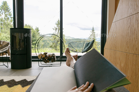 Beautiful female feet and book on background of fireplace and big window in modern chalet. Cozy home