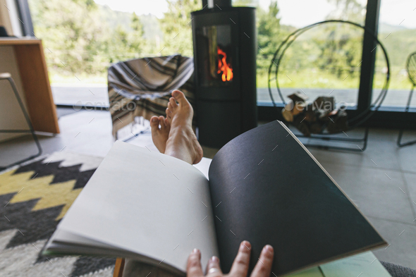 Beautiful female feet and book on background of warm fireplace and big window in modern cozy chalet